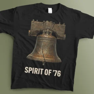 A flat lay photograph of a black graphic t-shirt laid flat on a muted green surface. The shirt features a highly detailed, vintage illustration of the Liberty Bell showing its iconic crack, rendered with oxidized copper and dark bronze metallic textures. Below the bell, the phrase "SPIRIT OF '76" is printed in a bold, weathered, off-white sans-serif font.
