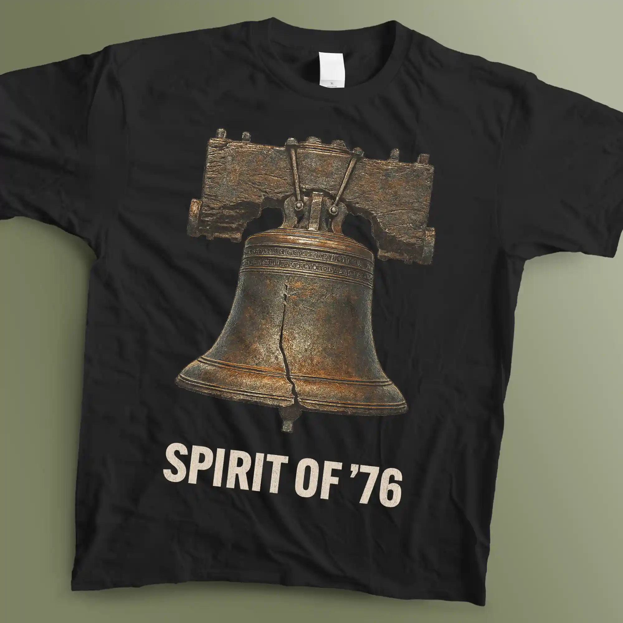 A flat lay photograph of a black graphic t-shirt laid flat on a muted green surface. The shirt features a highly detailed, vintage illustration of the Liberty Bell showing its iconic crack, rendered with oxidized copper and dark bronze metallic textures. Below the bell, the phrase "SPIRIT OF '76" is printed in a bold, weathered, off-white sans-serif font.